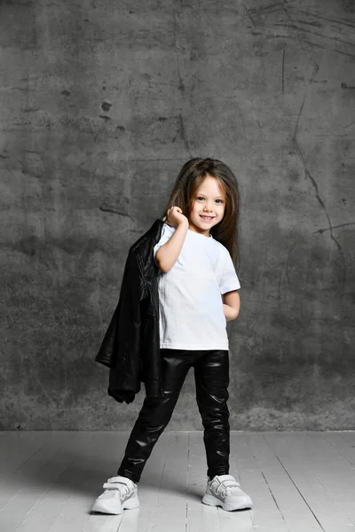 身穿黑白摇滚风格服装和白色运动鞋的积极向上的小女孩站在灰蒙蒙的混凝土背景上摆姿势 — 图库照片