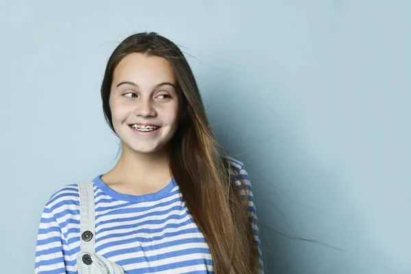 Tiener dame in jeans overall en gestreepte sweater. Ze glimlacht, kijkt opzij, poseert tegen een blauwe achtergrond. Sluiten. — Stockfoto