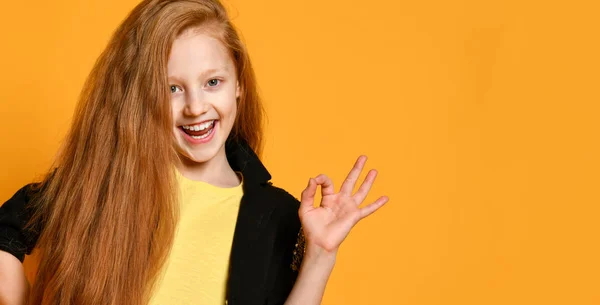 Ginger tienermeisje in zwarte jas, broek, geel t-shirt. Ze glimlacht, toont een goed teken, poseert op oranje achtergrond. Sluiten. — Stockfoto