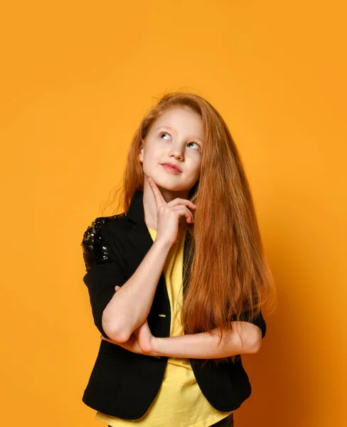 Ginger tienerkind in zwarte jas en broek, geel t-shirt. Ze ziet er attent uit, poserend op oranje achtergrond. Sluiten. — Stockfoto