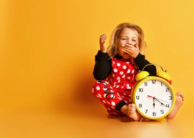 Küçük sarışın kız kırmızı noktalı ev kıyafetleri büyük sarı alarm saati ile yerde oturuyor ve sarı arka planda esniyor.
