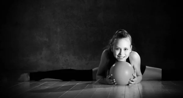 Giovane ragazza sorridente ginnasta in corpo nero sport e tomaie seduto in spago sul pavimento e tenendo la palla ginnica rosa in mano — Foto Stock