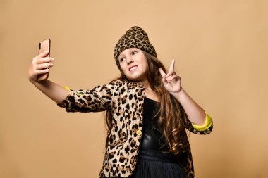Leopar renkli giysiler ve aksesuarlı genç kız ayakta duruyor ve akıllı telefonuyla selfie çekiyor ve barış işareti gösteriyor.