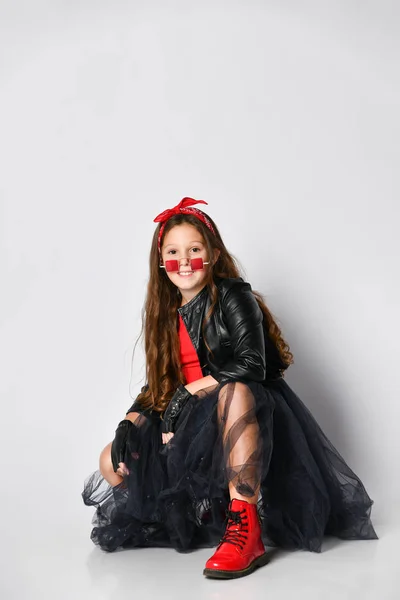 Joven chica sonriente en ropa de estilo rock brillante, botas rojas y accesorios sentados en el suelo con y mirando a la cámara — Foto de Stock