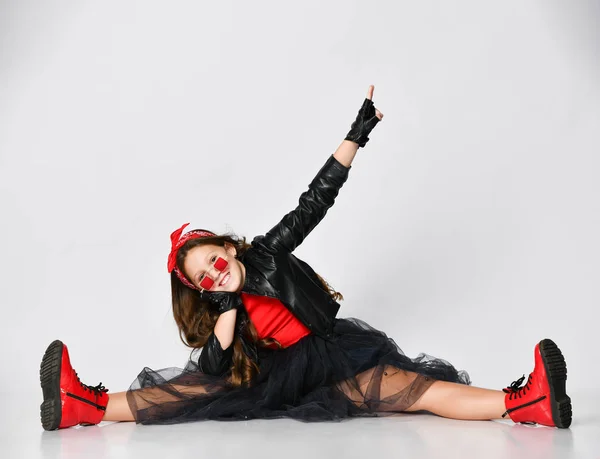 Joven chica positiva en ropa de estilo rock brillante y botas rojas sentadas en el suelo con las piernas estiradas y mostrando signo de paz — Foto de Stock