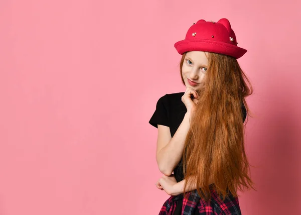 Adolescente in abito nero, camicia a scacchi in vita, cappello rosso. Sorride, si tocca il viso, posa su uno sfondo rosa. Da vicino. — Foto Stock