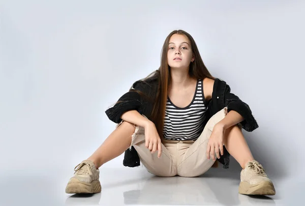 Mujer adolescente en camiseta a rayas, chaqueta vaquera negra, pantalones y zapatillas de deporte. Ella está sentada en el suelo aislada de blanco. De cerca. —  Fotos de Stock
