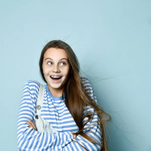 Teenage female in jeans overall, striped sweatshirt. She smiling, showing thumbs up, winking, posing on blue background. Close up — Stock Photo, Image