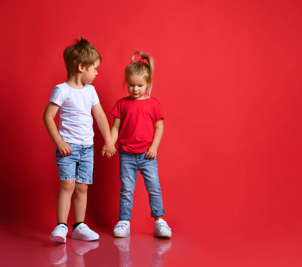 Small happy children boy and girl in stylish casual clothing standing, holding hands and feeling shy over red background