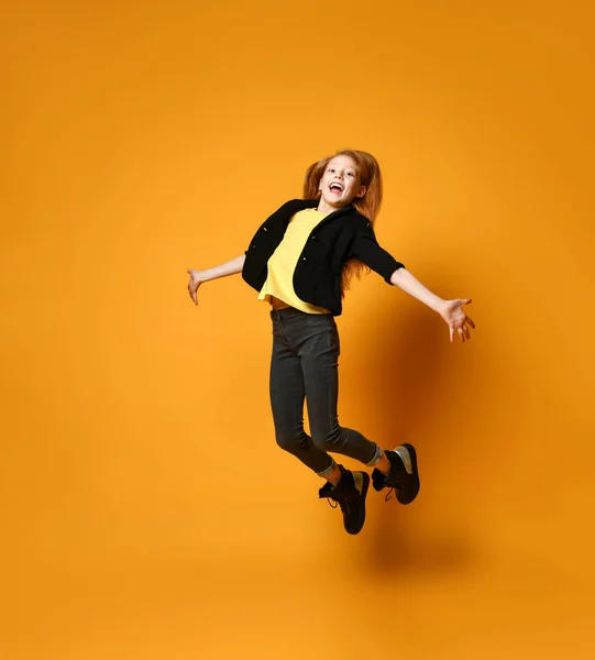 Teen female in black jacket, pants and boots, yellow t-shirt. She jumping up with spreaded hands, posing on orange background.