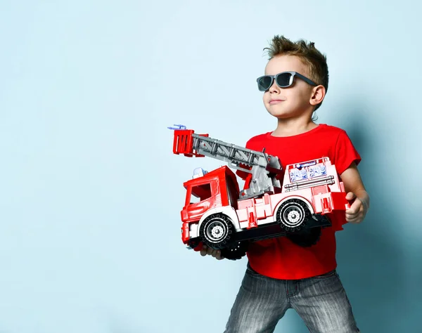 Positive boy in bright stylish casual clothing and sunglasses standing and holding toy fire engine in hands over blue background — ストック写真