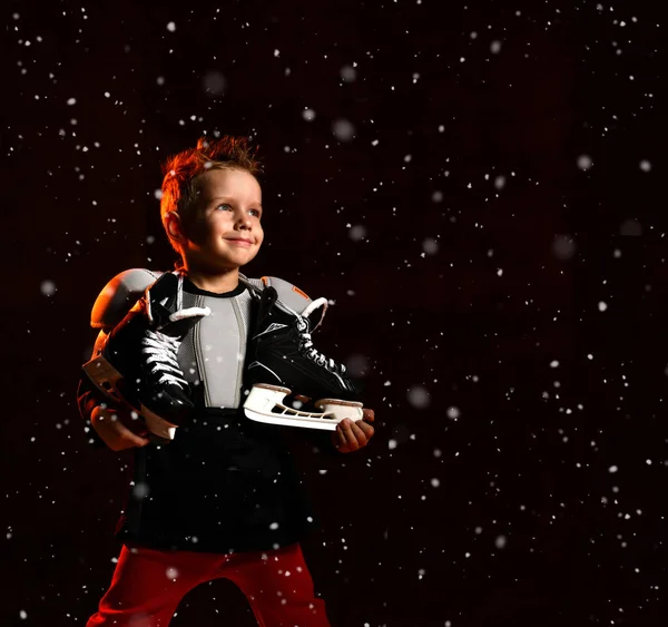 Niño rubio en uniforme de hockey con patines de hockey en el cuello de pie y mirando a la cámara sobre fondo oscuro — Foto de Stock
