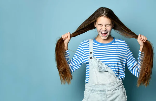 Adolescente en jeans en general y sudadera a rayas. Ella está gritando, sosteniendo su pelo largo, posando sobre fondo azul. De cerca. —  Fotos de Stock