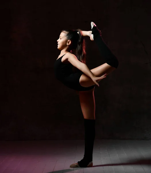Bambina in una ginnastica nera ginnasta body esegue un esercizio. Equilibrio su una gamba con una presa . — Foto Stock