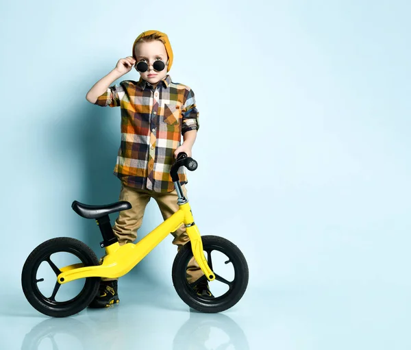 Jongen in heldere stijlvolle casual kleding, sneakers, pet en ronde zonnebril staan in de buurt van gele fiets aanwezig — Stockfoto