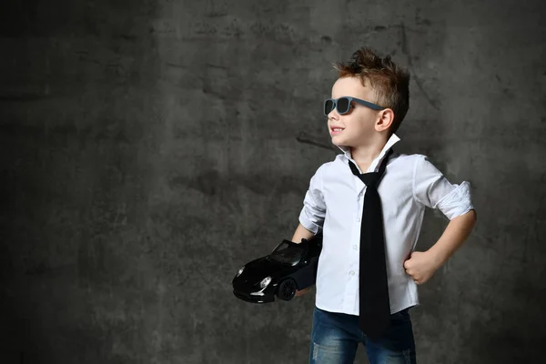 Uśmiechnięty chłopiec w stylowej, swobodnej białej koszuli, dżinsach, krawacie i okularach przeciwsłonecznych stojących i trzymających w ręku czarny samochodzik zabawki — Zdjęcie stockowe
