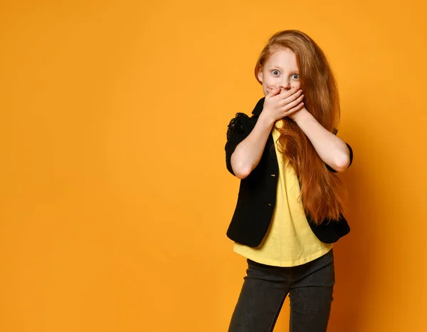 Teenage girl in black jacket and pants, yellow t-shirt. She is shocked, covered her mouth, posing on orange background. Close up — Stock Photo, Image