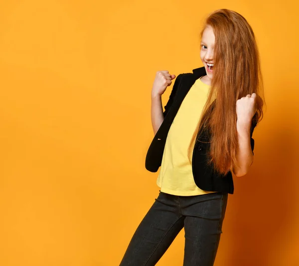 Ingefära skolflicka i svart jacka, byxor, gul t-shirt. Hon ler, ser nöjd ut, poserar på orange bakgrund. Närbild — Stockfoto