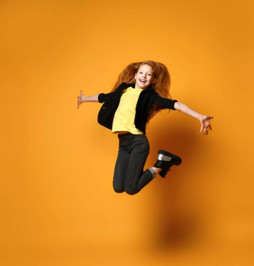 Siyah ceketli, pantolon ve botlu, sarı tişörtlü genç bir kadın. Kollarını açıp turuncu arka planda poz veriyor..