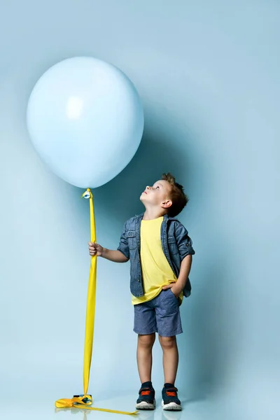 Şık giyinen sarışın çocuk, spor ayakkabıları mavi hava balonunun yanında duruyor ve yukarı bakıyor.