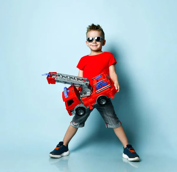 Pozitivní chlapec v jasném stylovém ležérní oblečení a sluneční brýle stojící a držící hračku požární motor v rukou přes modré pozadí — Stock fotografie
