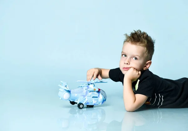 Pozytywne blond chłopiec w stylowej odzieży casual siedzi na podłodze i trzymając helikopter zabawka obecny w ręku nad niebieskim tle — Zdjęcie stockowe