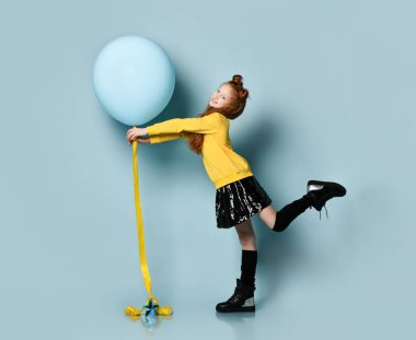 Sarı kazaklı, siyah etekli, diz boyu çizmeli bir çocuk. Gülümsüyor, balonu tutuyor, mavi arka planda yan poz veriyor..