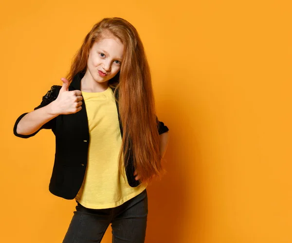 Rödhårig skolflicka i svart jacka och byxor, gul t-shirt. Hon visar tummen upp, poserar på orange bakgrund. Närbild — Stockfoto