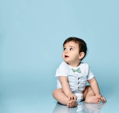 Beyaz tulumlu, papyonlu, yalın ayaklı bir bebek. O, mavi arka planda yerde oturuyor. Kapat.