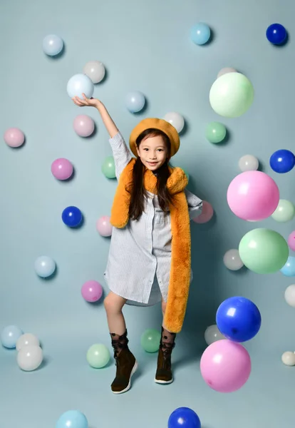 Little asian kid in oversized shirt dress, brown beret and scarf, boots. She smiling, posing with balloons on blue background.