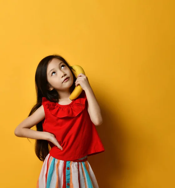 Aziatisch kind in rode blouse, veelkleurige rok. Ze deed alsof ze op banaan praatte als per telefoon, poserend op oranje achtergrond.. — Stockfoto