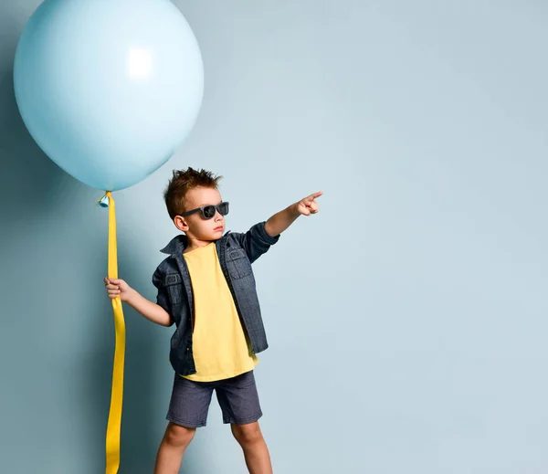 Blonďatý chlapec ve stylovém neformálním oblečení, teniskách a slunečních brýlích stojící poblíž modrého vzduchového balónu a ukazující vpravo — Stock fotografie