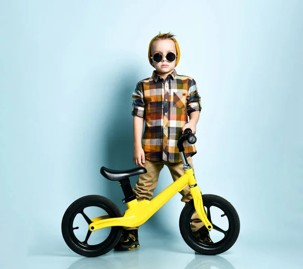 Pozytywny chłopiec w jaskrawo stylowej odzieży casual, trampki i czapki siedzi na żółty rower bieg obecny na niebieskim tle — Zdjęcie stockowe