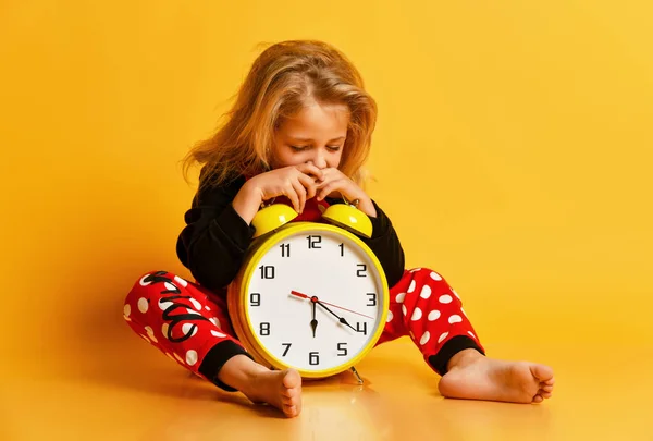 Malá blondýnka v červených tečkovaných pyžamech sedící na podlaze s velkými budíky, unavená, zívající a spící — Stock fotografie