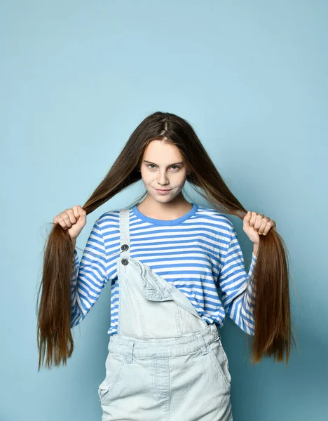 Tini nő farmerben és csíkos pulóverben. Hosszú haját fogja, kék stúdió háttérrel pózol. Közelről. — Stock Fotó