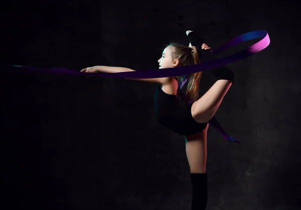 Молодая девушка гимнастка в черном спортивном теле и верхние стоят боком и делают упражнения с синей гимнастической лентой — стоковое фото