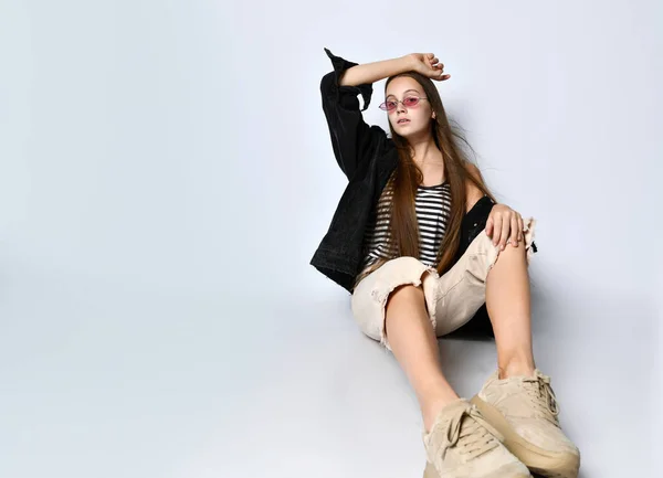 Tonårstjej i solglasögon, randig t-shirt, svart jeansjacka, byxor och sneakers. Hon satt på golvet isolerad på vitt. Närbild — Stockfoto