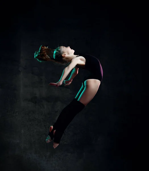 Giovane ginnasta ragazza in corpo nero sport e tomaie speciali saltare in posa ginnica su sfondo scuro — Foto Stock
