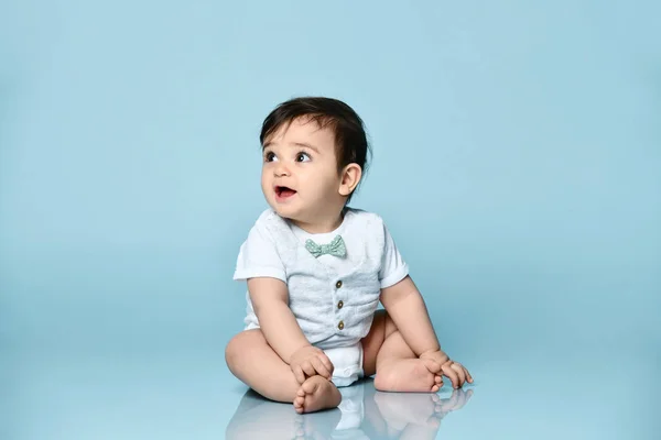 Petit bambin en body blanc en gilet avec noeud papillon, pieds nus. Il est assis sur le sol sur fond bleu. Gros plan — Photo