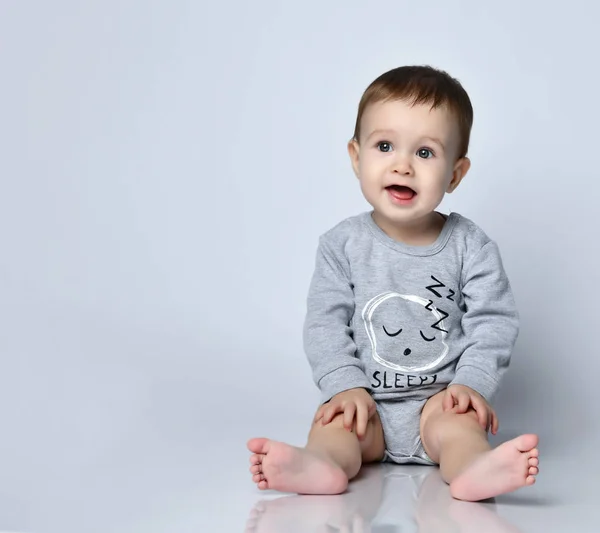 Bambino bambino in tuta casual grigio e a piedi nudi seduto sul pavimento e sorridente su sfondo bianco parete — Foto Stock