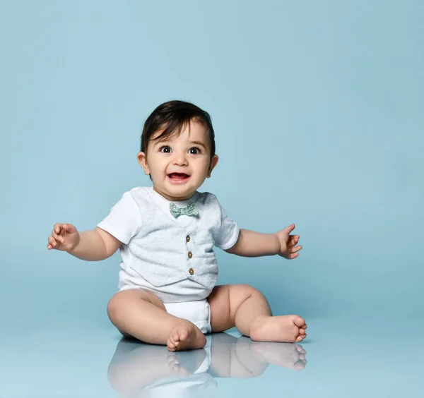 Bébé garçon en body blanc en gilet avec noeud papillon, pieds nus. Il sourit, assis par terre sur fond bleu. Gros plan — Photo