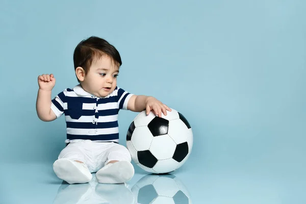Niño en camiseta a rayas, pantalones blancos y botines. Está mirando hacia arriba, sentado en el suelo sobre fondo azul. De cerca. —  Fotos de Stock