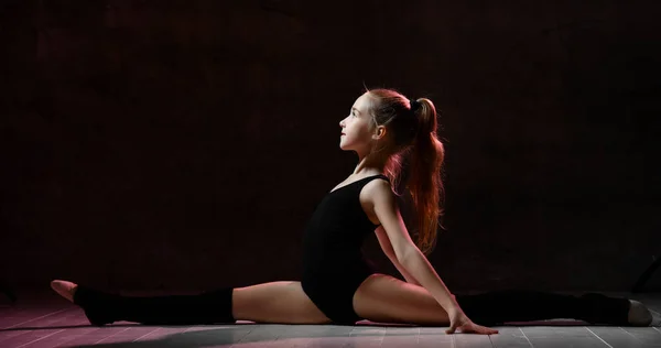 Giovane ragazza sorridente ginnasta con i capelli lunghi in corpo nero sport e tomaie seduti in spago lateralmente su sfondo scuro — Foto Stock