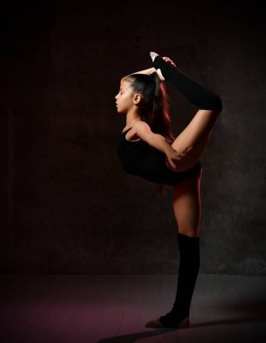 Siyah jimnastik mayosu giymiş küçük bir kız egzersiz yapıyor. Bir bacağa tutunarak denge.