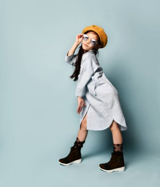 Güneş gözlüklü, büyük beden gömlekli, kahverengi bereli, çizmeli Asyalı bir çocuk. Seni öpüyor, mavi arka planda poz veriyor. Tam uzunluk