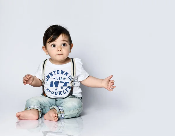 Bambino in t-shirt con iscrizioni, jeans con bretelle. È seduto sul pavimento isolato su sfondo bianco. Da vicino. — Foto Stock