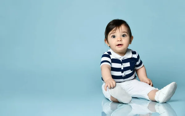 Bébé garçon en t-shirt rayé, pantalon blanc et chaussons. Il sourit, assis sur le sol sur fond bleu studio. Gros plan — Photo