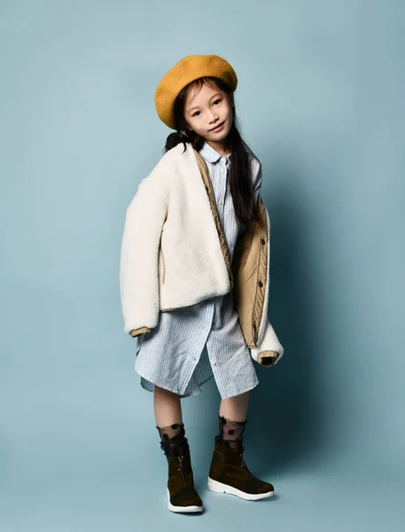 Petite écolière asiatique en robe chemise, veste double face, béret marron, bottes. Souriant, posant sur fond bleu. Longueur totale — Photo