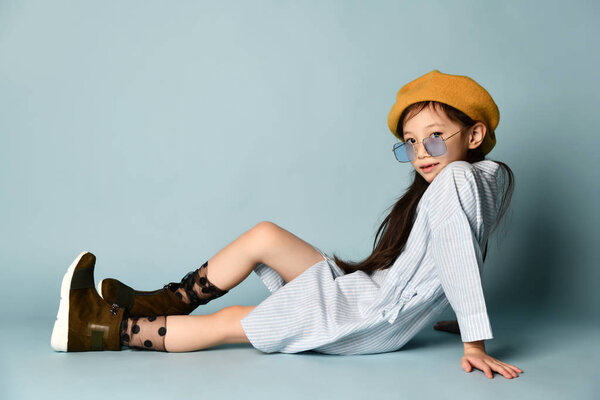 Маленький азиатский мальчик в солнечных очках, в рубашке, в коричневом берете, в сапогах. Улыбаясь, позируя сидя боком на синем фоне. Закрыть
