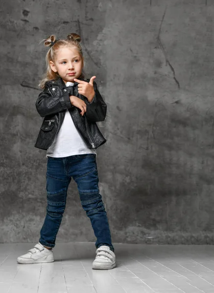 Klein schattig lachend blond meisje in stijlvolle rock stijl zwart lederen jas, jeans en witte sneakers staan in zelfverzekerde pose — Stockfoto